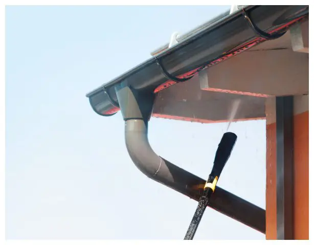 Pressure washing house gutters under clear sky.